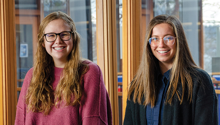 2 female college students. The left student is wearing plum knit sweater with dark rimmed glasses. The other student is wearing a dark blue sweater with metal rimmed glasses.