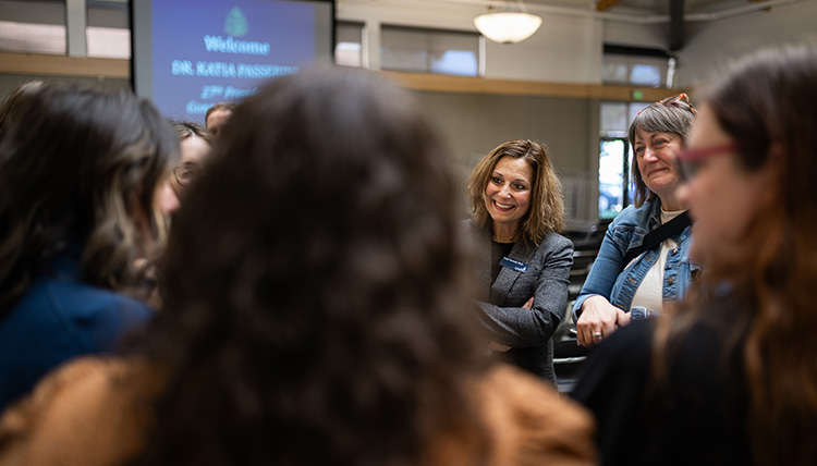 A group of people standing in a circle, conversing in front of a screen that reads 'Welcome DR. KATIA PASSERINI' in an indoor event space with large windows and overhead lighting.