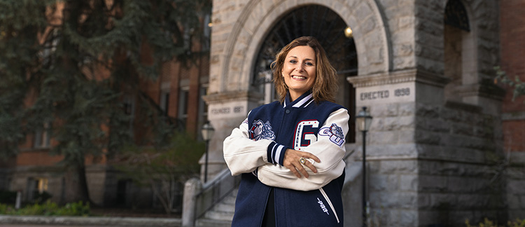 A person stands in front of a historic stone building with an arched entrance wearing a navy blue and white varsity jacket with patches on the sleeves and chest, including a large 'G' on the right side.