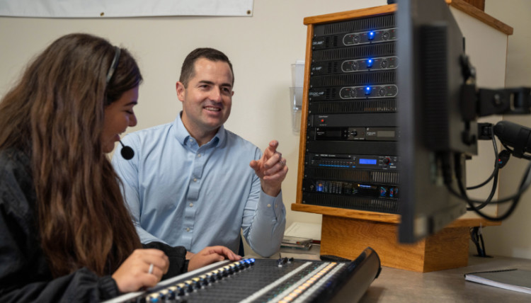 Broadcast journalism student operating an audio mixing console while a faculty member provides guidance in a university media control room equipped with professional sound and production equipment.