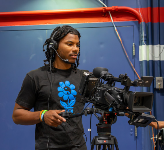Broadcast journalism student operating a professional video camera with headphones in a university studio, learning camera techniques and live video production.