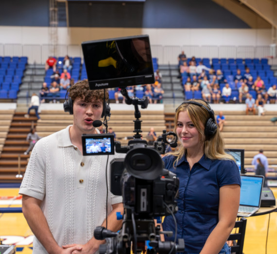 Broadcast journalism students operating a professional video camera with monitor and wearing headsets during a live sports event broadcast in a university gymnasium.