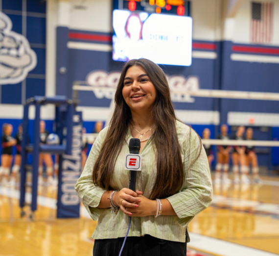 Broadcast journalism student reporting from a ������ University volleyball game, holding an ESPN microphone on the court with players and scoreboard visible in the background.
