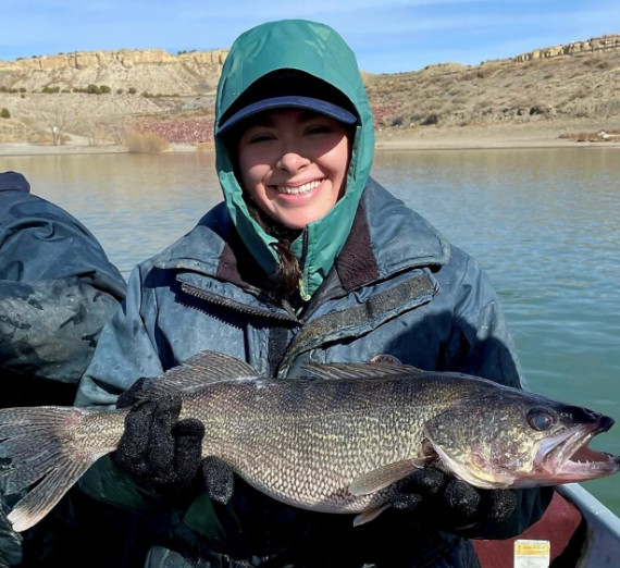 Alumni Jordan Cruz holding a large Walleye fish in front of a body of water. 
