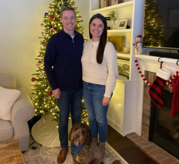 A man and woman standing in front of a Christmas tree with a shaggy brown good dog sitting in front of them. 