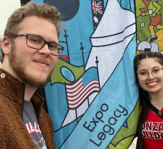 Two Gonzaga History students stand next to a colorful Expo ’74 Legacy banner at Spokane’s Riverfront Park during the 50th anniversary celebration. Banner features iconic fair imagery including flags and pavilion design, highlighting the History Department’s donor-supported Reader’s Theatre project.