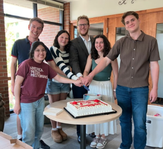 Group of 91 History Department students gathered around a table cutting a celebratory cake during a Phi Alpha Theta event. One student wears a maroon T-shirt with the text ‘America Needs Historians.’