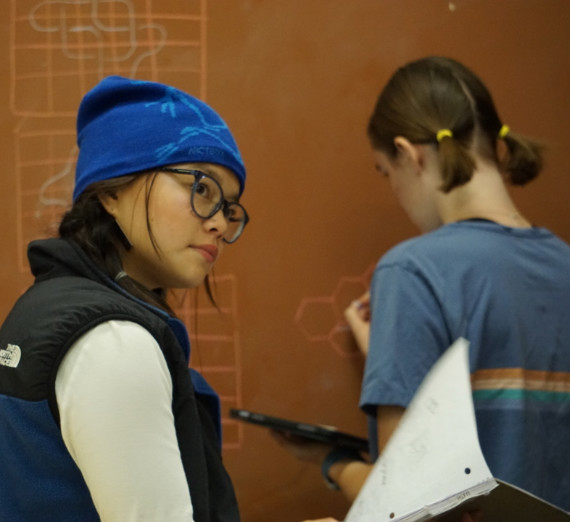 Two students in a math classroom-one holding a notebook and listening, one writing on the board. 
