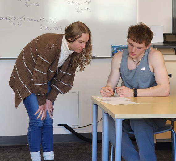 A student sitting at a table with work in front of him, and a tutor leaning over to look. 