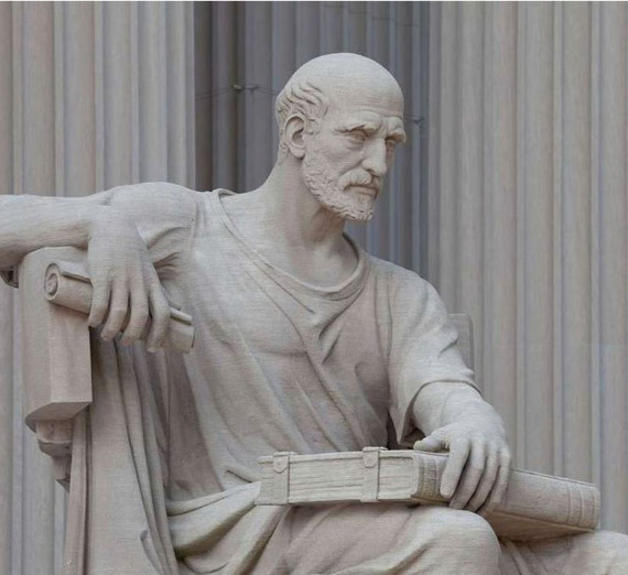 Statue of a seated man in ancient Greek or Roman style clothing holding a scroll in one hand with his other hand over a book in his lap. 
