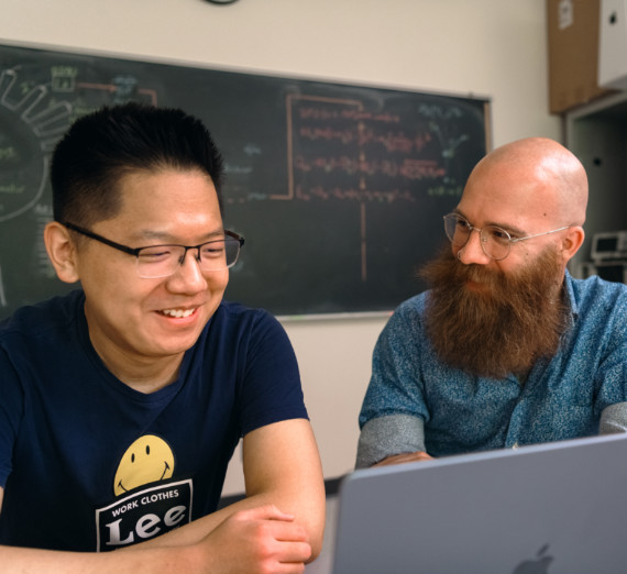 Dr. Adam Fritsch and a student in a classroom smiling together while looking at a computer. 