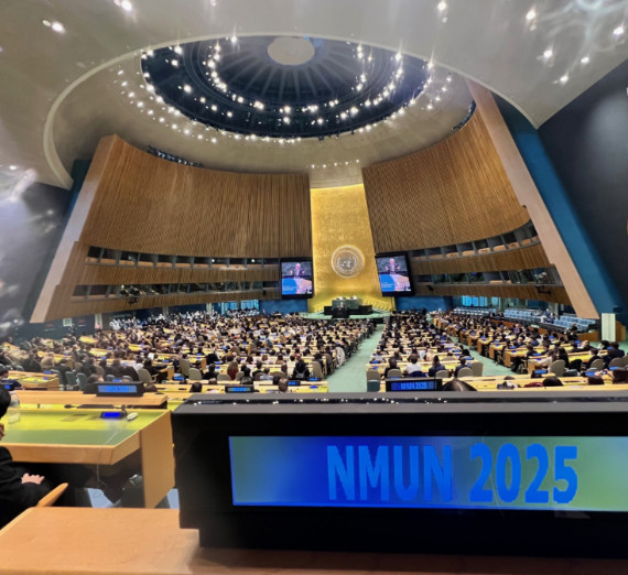 National Model United Nations 2025 conference inside the United Nations General Assembly Hall in New York City. The photo shows hundreds of delegates seated in rows, the iconic gold UN emblem on the wall, large screens displaying proceedings, and the modern circular ceiling design.