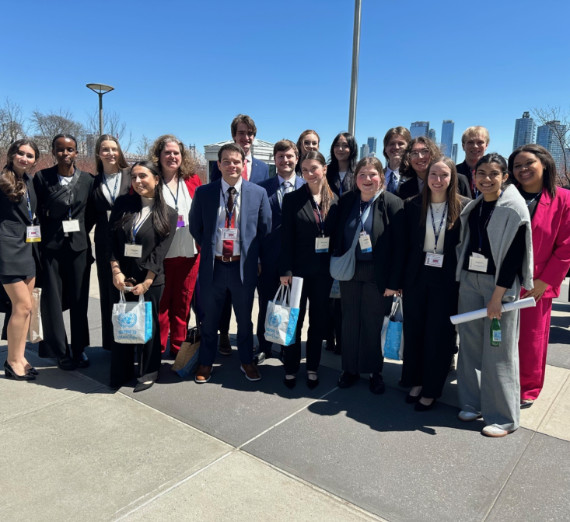Gonzaga University Model United Nations delegation posing outside the United Nations Headquarters in New York City during the spring conference. Students are dressed in professional business attire, wearing conference badges and holding UN-branded tote bags, with a clear blue sky and city skyline in the background.