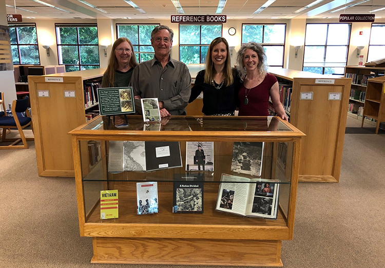 Group photo of Larry Plager and the Telling War group gathered around a bookcase with a collection of Vietnam war books.