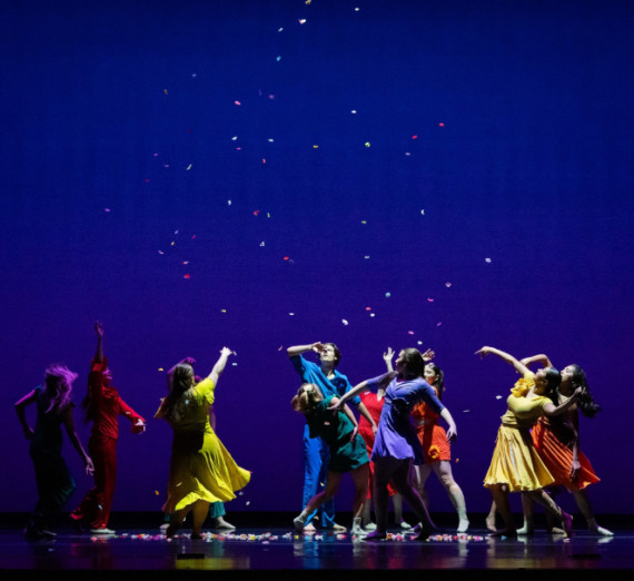 A group dancing in front of a blue background in bright primary color costumes.