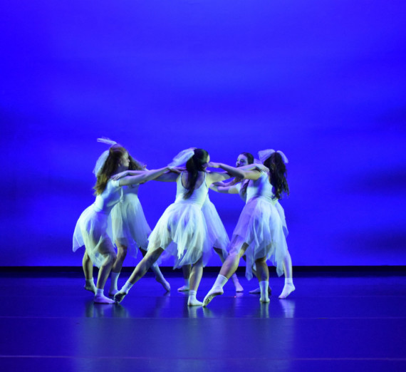 A group of dancers in white dresses dancing in a circle against a blue background. 
