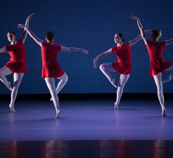 Four dancers in red dresses and white tights dancing in a cirle with arms in the air, against a blue background.