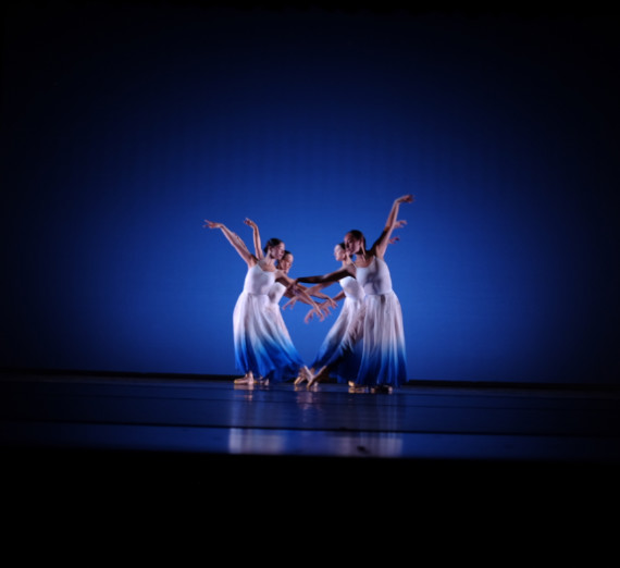 Four dancers in dresses on stage with a blue background. They are in two rows of two with center arms and feet pointing toward the ground, and their outer arm pointing up.