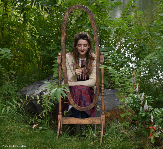 A woman sitting in front of a river looking down at a leaf in her hand. She is wearing a skirt and top that look like they're possibly from the 1950's and there is an empty mirror frame in front of her, framing her. 