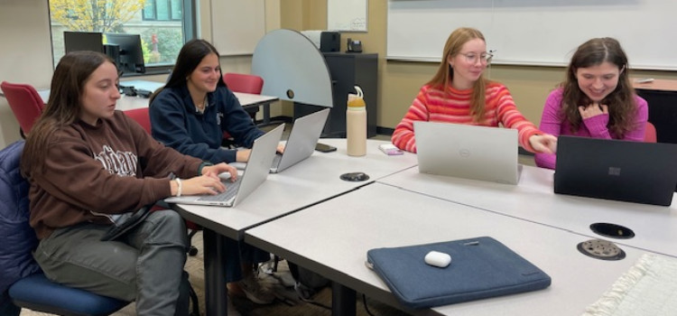 Four students working together on laptops around a conference-style table in a modern classroom with whiteboards, a projector screen, and large windows. A tablet and water bottle are on the table, highlighting a collaborative learning environment with digital tools.