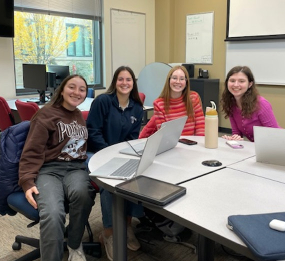 Students collaborating in a modern classroom with laptops and tablets on a round table, engaged in a group project. The room features large windows, whiteboards, and a projector screen, creating an interactive learning environment.