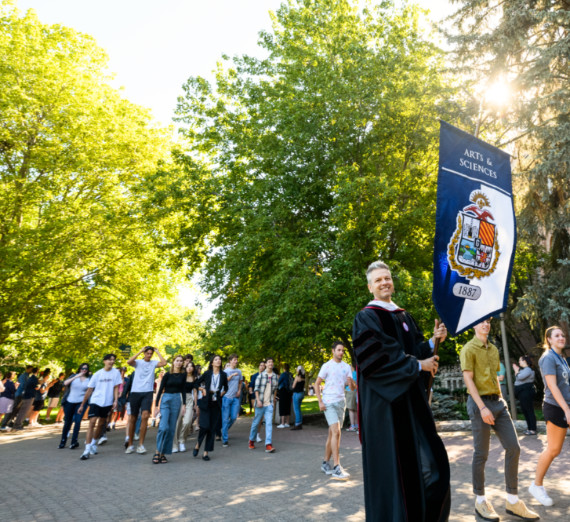 Faculty member in regalia holding a Banner in a procession with students on a tree lined path.