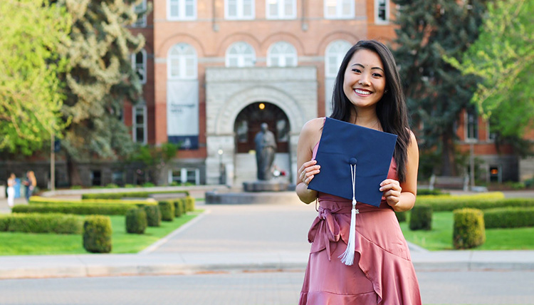 Gonzaga Alumni Allana Platt stands in front of College Hall holding a graduation cap.