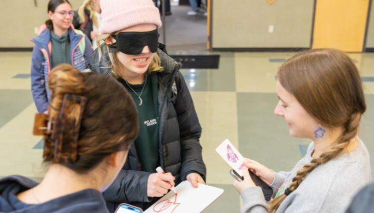 Two girls describing a picture to a blindfolded person who is trying to draw what they see