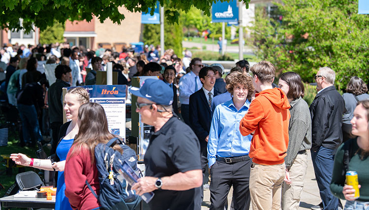 A crowd of people of all ages gather outside on a sunny day along sidewalks and tables of academic posters, talking and pointing to machines, computers and photographs.