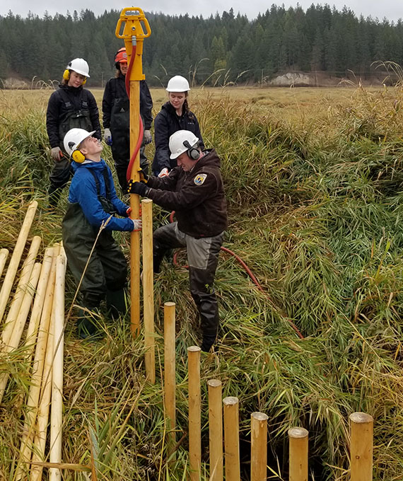 Four students in hard hats and coveralls hold a tall pole steady in a stream. More poles stand erect across the stream.