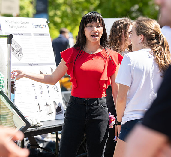 A young female student talks to another woman while pointing at an academic poster outside on a sunny day
