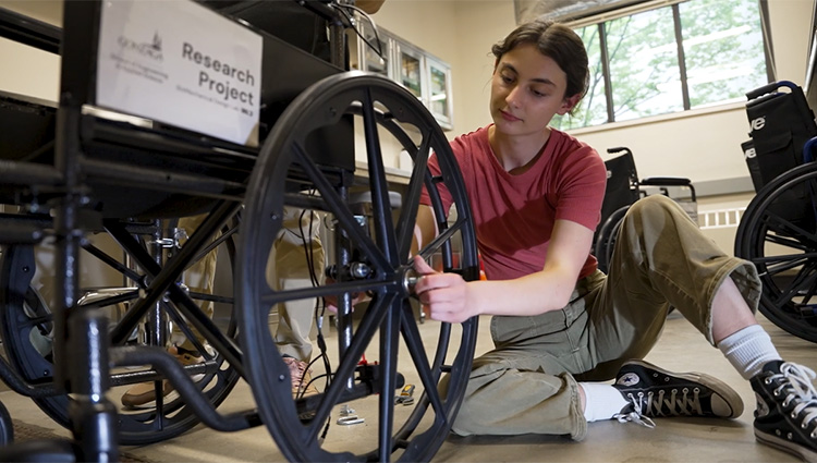 A young person in a red t-shirt and olive pants sits on the floor, adjusting the wheel of a black wheelchair. A "Research Project" sign is visible on the chair.