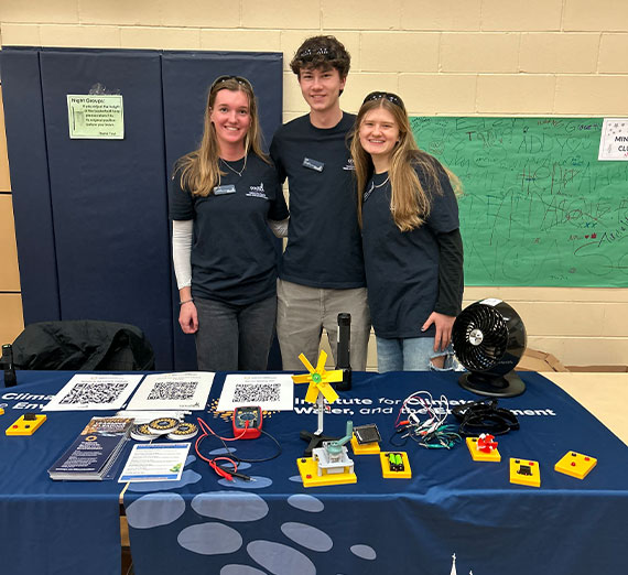 Three young adults stand smiling behind a table at an indoor event, all wearing matching dark blue shirts and name tags. The table in front of them displays several printed QR codes, a small yellow wind turbine model, and a black fan. Behind them, a padded gym wall and a green poster with various handwritten messages are visible.