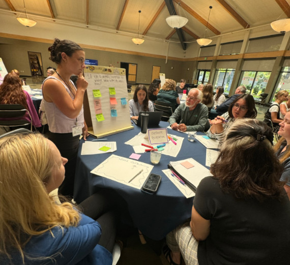 A facilitator stands at a round table listening to a smoke symposium participant share feedback