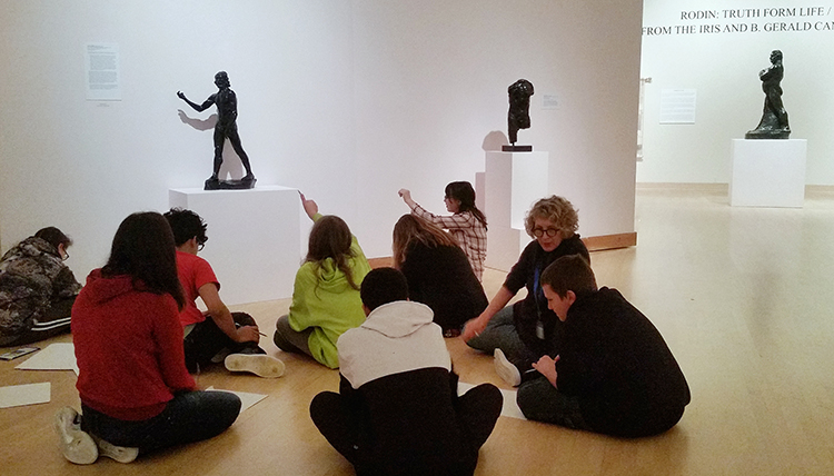 Middle school students sit on the floor of the museum discussing bronze art  on display.