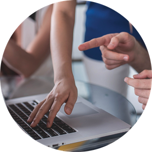 A laptop sits on a rounded glass table, in front of three people out of the screen. The hands of the rightmost person points towards the screen, while the person in the middle reaches down to type with one hand on the keyboard. A third person is visible to the left with elbows resting on the same table.