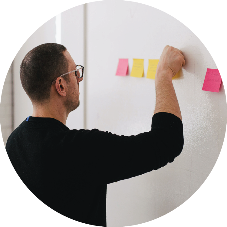 A man in a black sweater stands in front of a whiteboard and reaches up to place a colored sticky note alongside a line of yellow and pink notes.