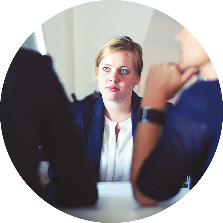 A young woman wearing a blazer sits across from two people, whose backs are to the camera. She appears to listen intently to one of the people across from her.