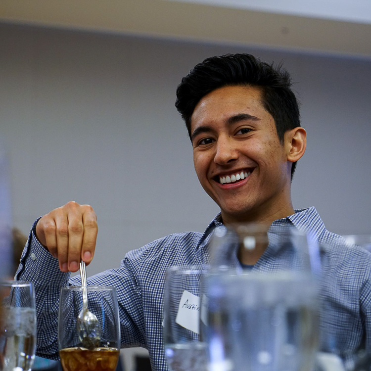 A male student wearing a blue button down shirt smiles at the camera as he stirs a spoon in a glass of iced tea.