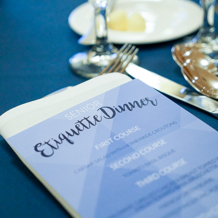 A printed menu rests on a table with a dark blue tablecloth. The menu header reads 'Senior Etiquette Dinner' with the first, second, and third course listed. Silverware is visible just to the left of the menu as well as two stems from wine-style glasses.