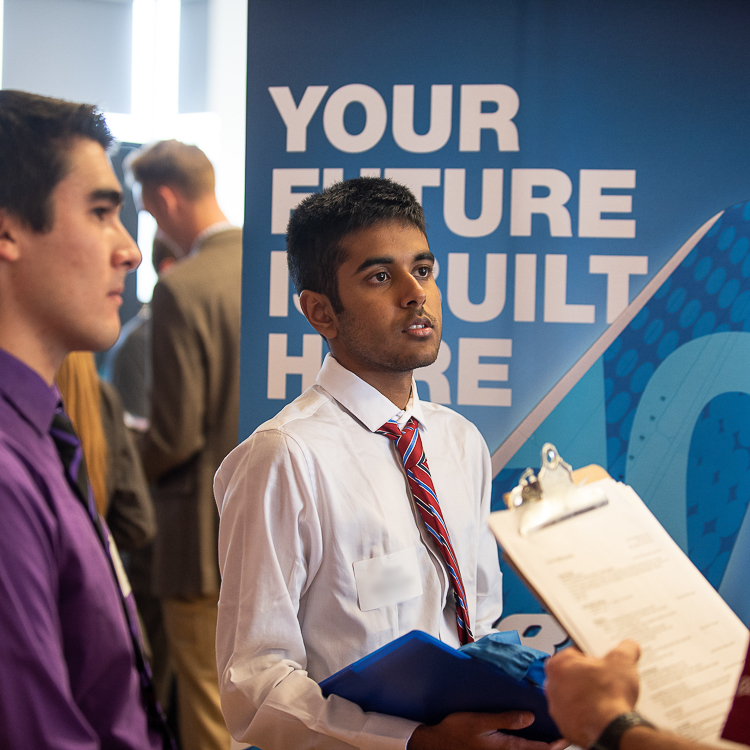 A young man wearing a white dress shirt and red tie listens to an employer representative at the All Majors Career Fair.