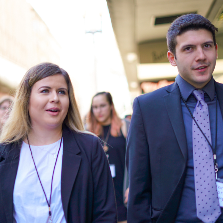 Two students walk side-by-side on the San Francisco trek. The each wear a formal blazer and a lanyard with their name tag on it.