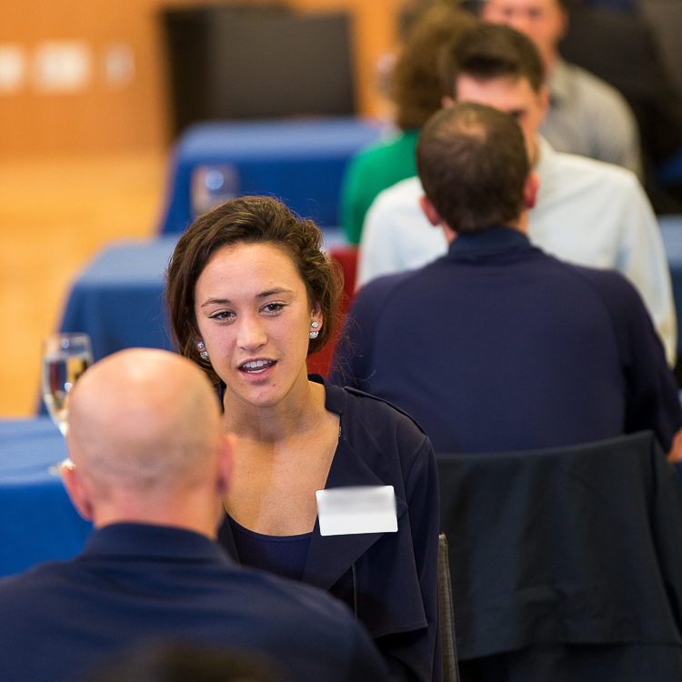 A young woman practices her interview answer with a mock interviewer sitting opposite her at a table. Visible behind her are two more tables in the row with students and mock interviewers similarly interacting.