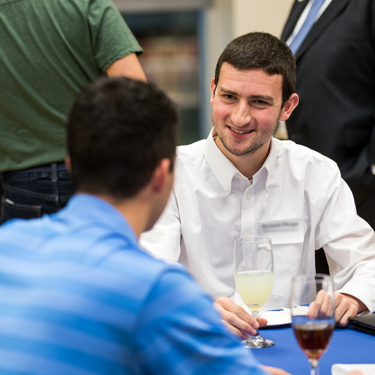 A young man in a white dress shirt smiles as he listens to another person with their back to the camera wearing a blue dress shirt.