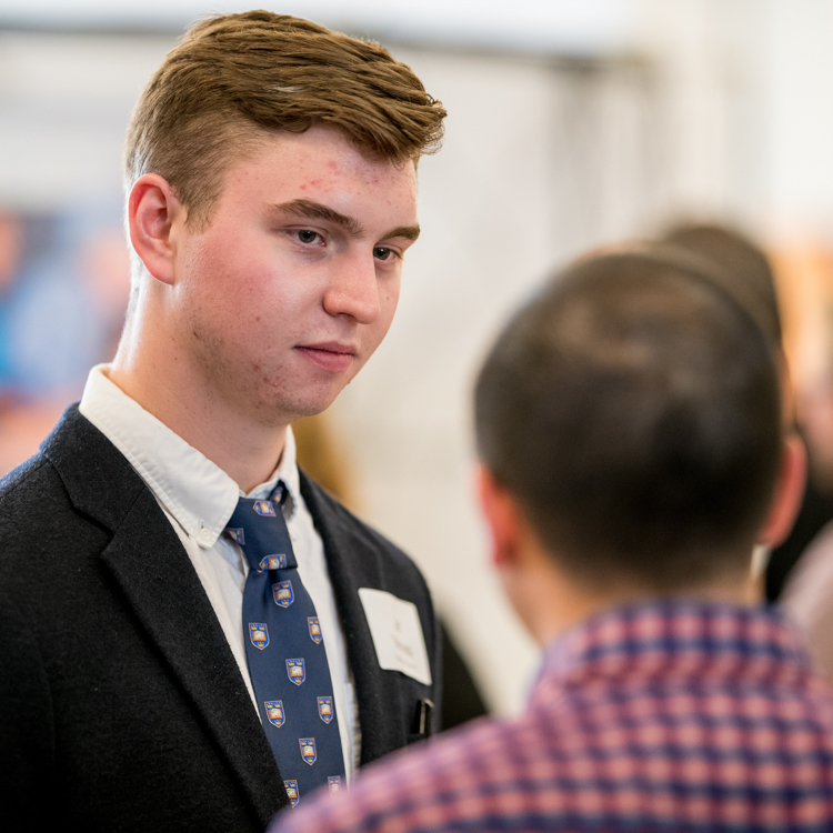 A young man in a formal blazer and tie listens to a recruiter in front of him. The recruiter has his back to camera.