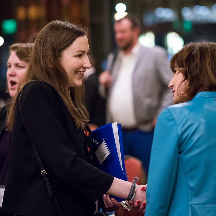 A young woman in a black blazer shakes the hand of an alumni wearing a blue jacket.