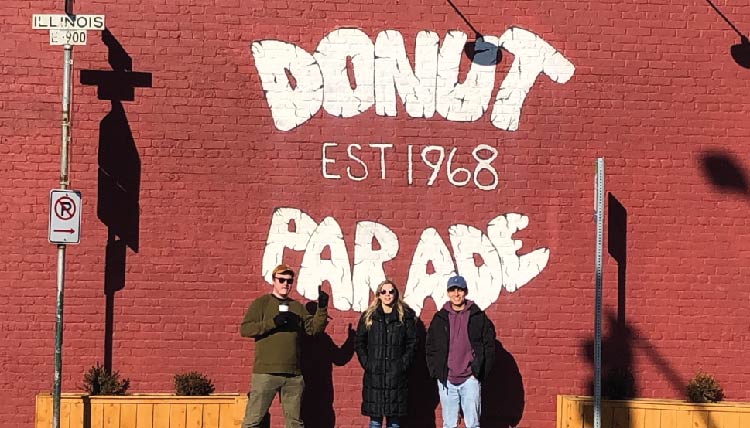 three students standing in front of a building with the words 'Donut Parade, est 1968' inscribed on it
