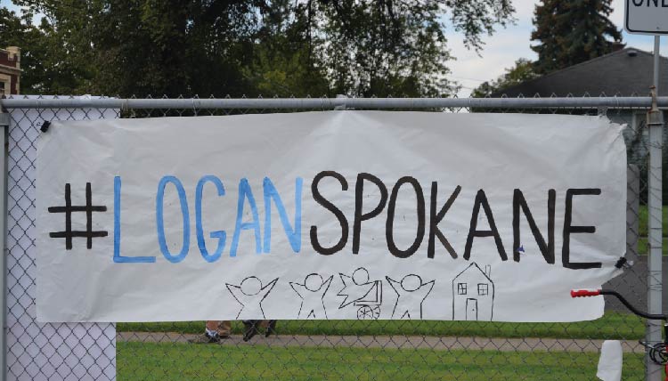 a hand-drawn sign fastened to a chain-link fence. The sign reads #LoganSpokane with a illustration of children and a house on the bottom of the sign.