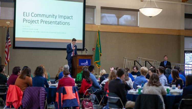 Student presenter in a conference setting with the words, "ELI Community Impact Project Presentations" on a background screen.