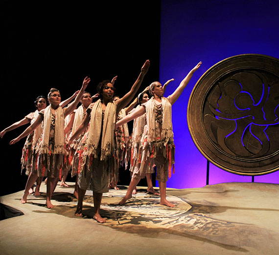 Performers on stage in fringed costumes, extending arms in a choreographed dance under purple lighting.